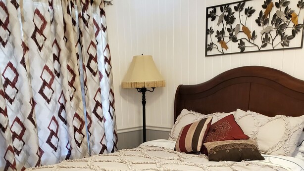 Una cama bien hecha con una colcha blanca con volantes y varios almohadones, un cabecero de madera oscura, una lámpara de pie y una pared con un adorno metálico.