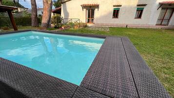A rectangular swimming pool with a dark textured edge, surrounded by a grassy lawn and a building with a tiled roof and green shutters.