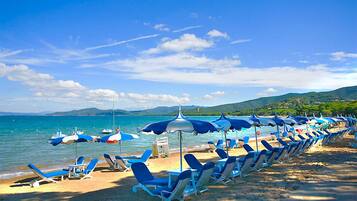 Una spiaggia con file di sdraio blu e ombrelloni abbinati, un mare cristallino e un paesaggio collinare sullo sfondo.