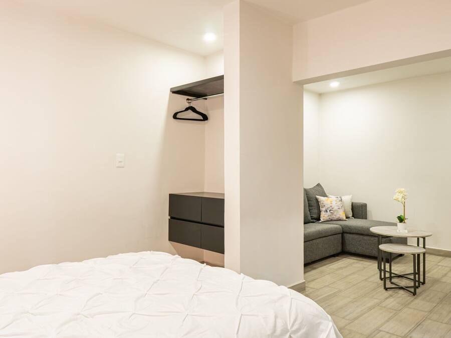 A modern bedroom with a large bed, a small bedside table, a grey sofa, and a round table with a vase of flowers.
