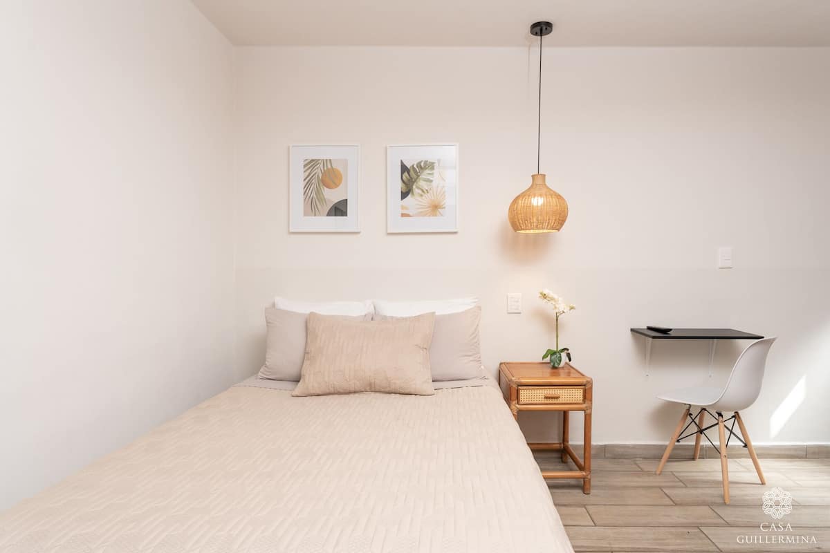 A bedroom with a bed, bedside table, chair, desk, and two framed artworks.