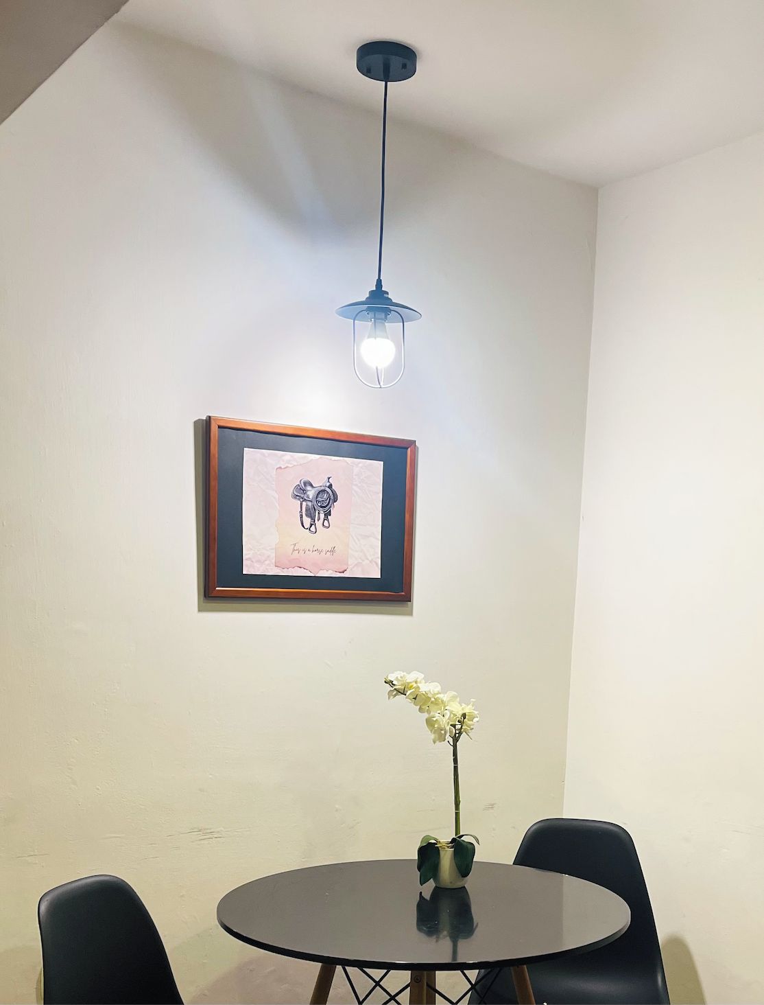 Un coin repas comprenant une table ronde, deux chaises, un tableau encadré au mur et un luminaire suspendu.