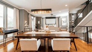 A spacious dining area with a long wooden table, two white cushioned chairs, a chandelier, and a staircase in the background. - Midtown Houston Townhome - Minutes from downtown and night life! (Houston)