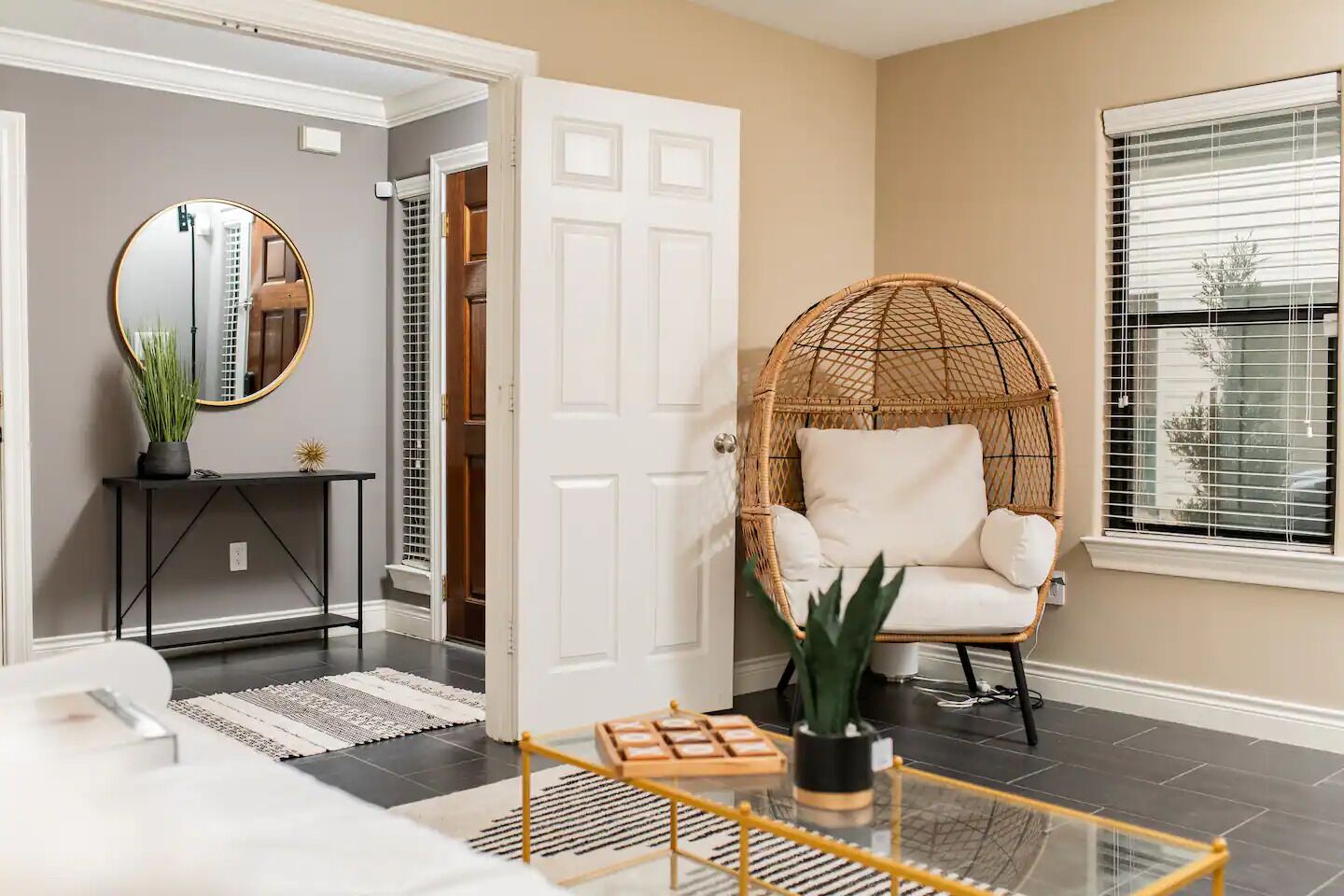 A modern living room with a wicker chair, a round mirror, a console table, and a potted plant.