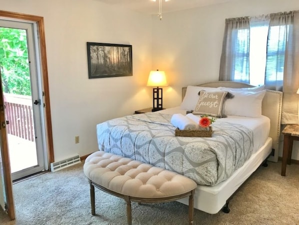 A bedroom with a bed, a bench, a nightstand, a lamp, a picture on the wall, and a window with curtains.