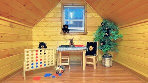 A cozy attic room with wooden walls, a small window, a crib, a toy table, and a Christmas tree.