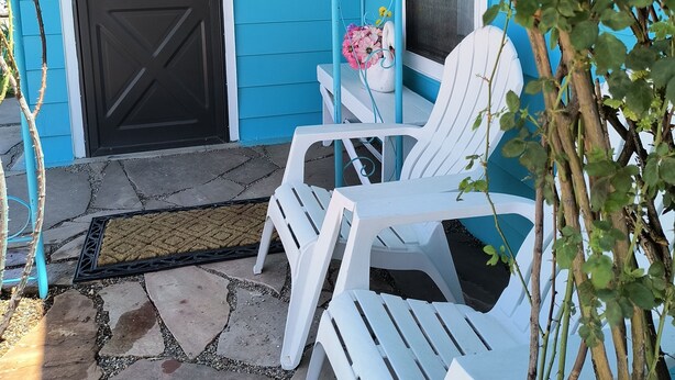 Une maison bleue avec une porte noire, des chaises en plastique blanches et une petite table sur laquelle est posé un vase de fleurs.