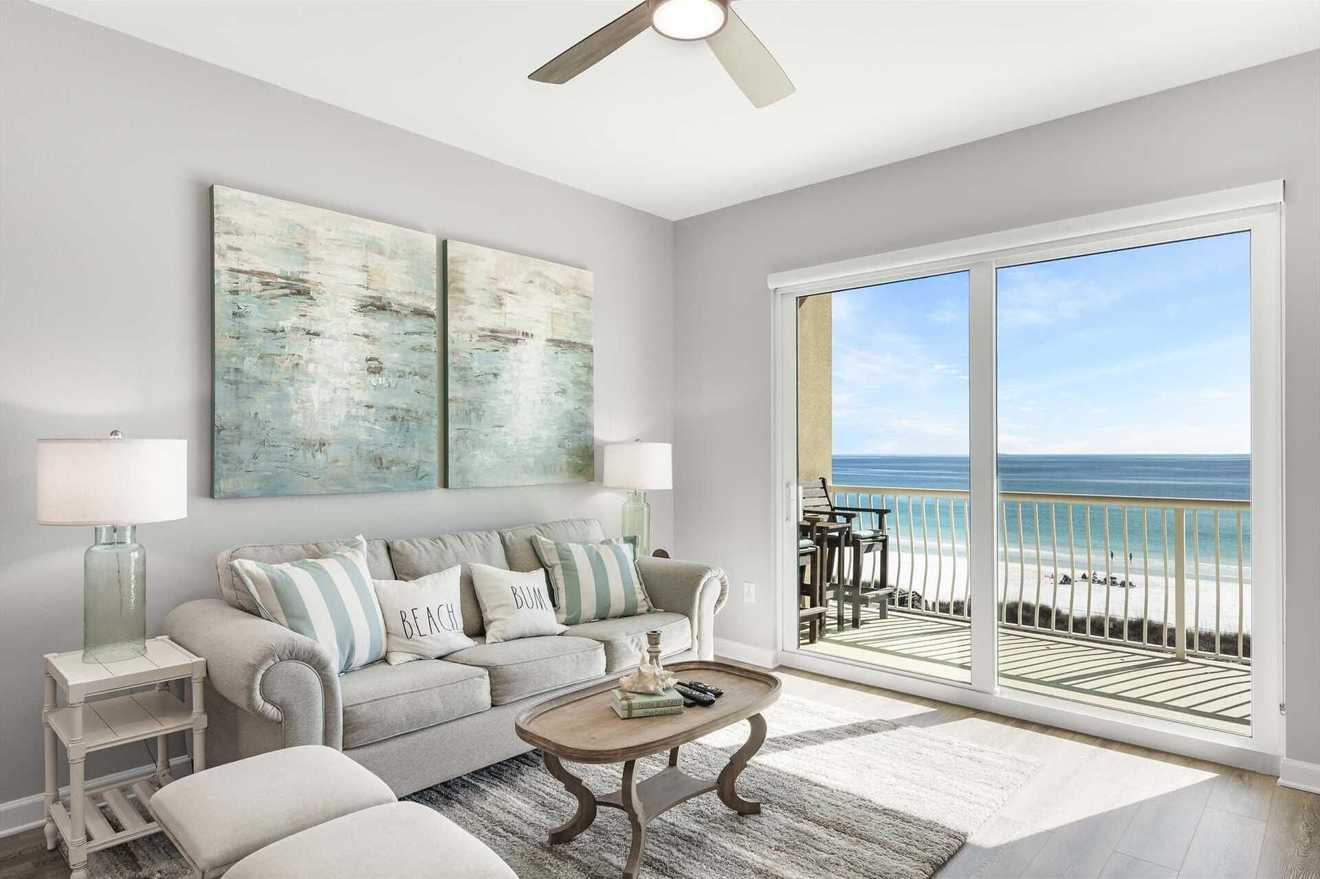 A living room with a large window overlooking a beach, a ceiling fan, and a sofa with beach-themed pillows.