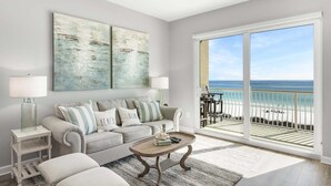 Ein Wohnzimmer mit großem Fenster zum Strand, Deckenventilator und einer Couch mit Strandkissen.