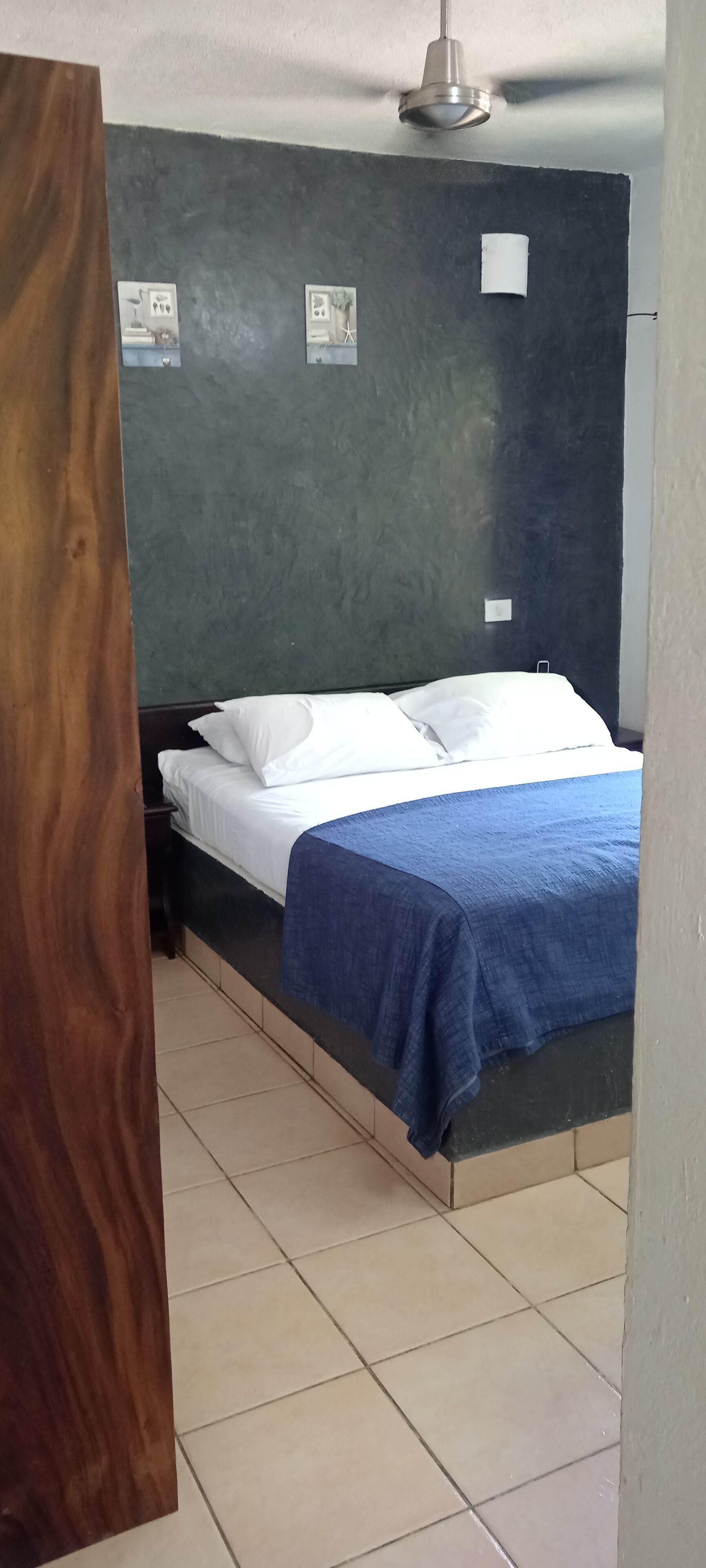 Un dormitorio con una cama, una mesita de noche, un ventilador de techo y una pared con dos ventanas pequeñas.