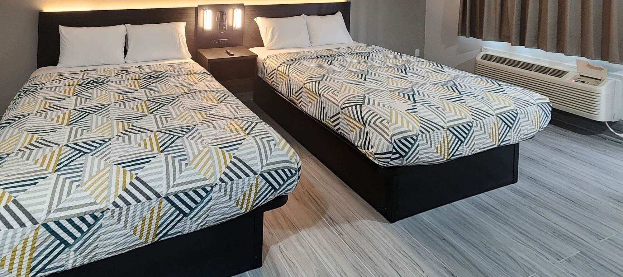 A hotel room with two beds, each with a geometric-patterned bedspread, a nightstand with a remote control, and a wall-mounted heater.