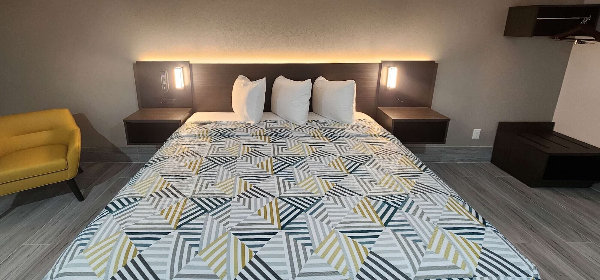 Una camera d'albergo con un letto grande, due comodini con lampade, una poltrona gialla e una coperta moderna con motivi geometrici.