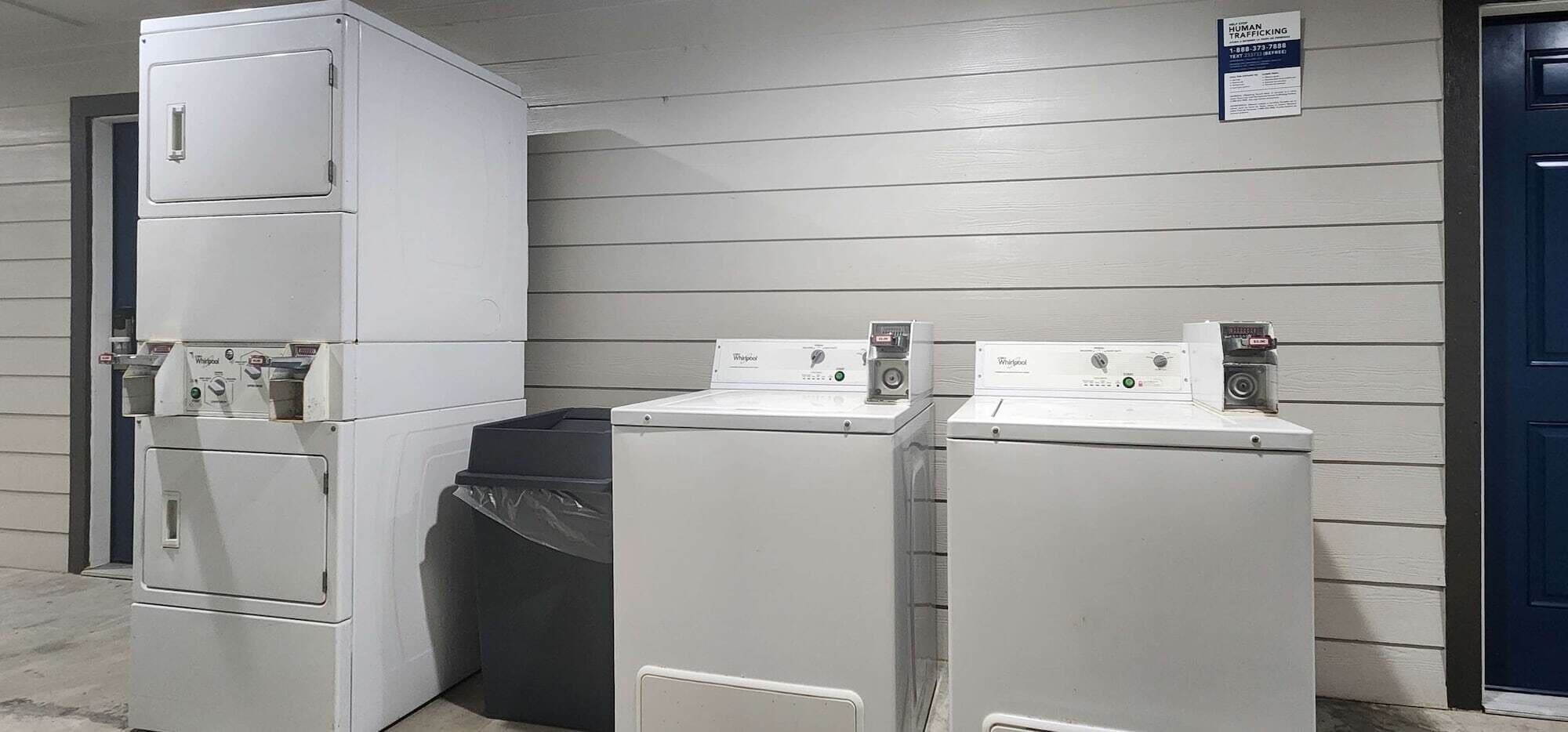 Un área de lavandería con lavadoras y secadoras apiladas, un contenedor de basura y una puerta con un letrero.