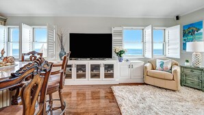 A dining area with a wooden table and chairs, a television on a cabinet, and a view of the ocean through open windows. - Spectacular direct ocean front condo with amazing views of the Atlantic (Satellite Beach)