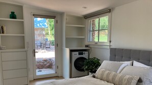 A bedroom with a bed, a washing machine, a window, and a door leading to an outdoor area.