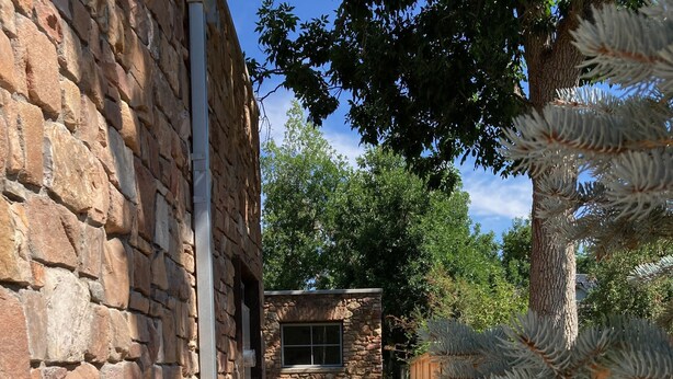 A gravel pathway leading to a stone building with a window, surrounded by greenery and a wooden fence.
