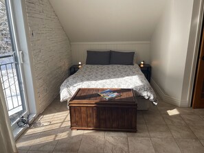 Ein Schlafzimmer mit schrägem Dach, einem Bett mit floralem Überwurf, zwei Nachttischen mit Lampen, einer hölzernen Kommode und einem Fenster mit Blick auf Bäume.
