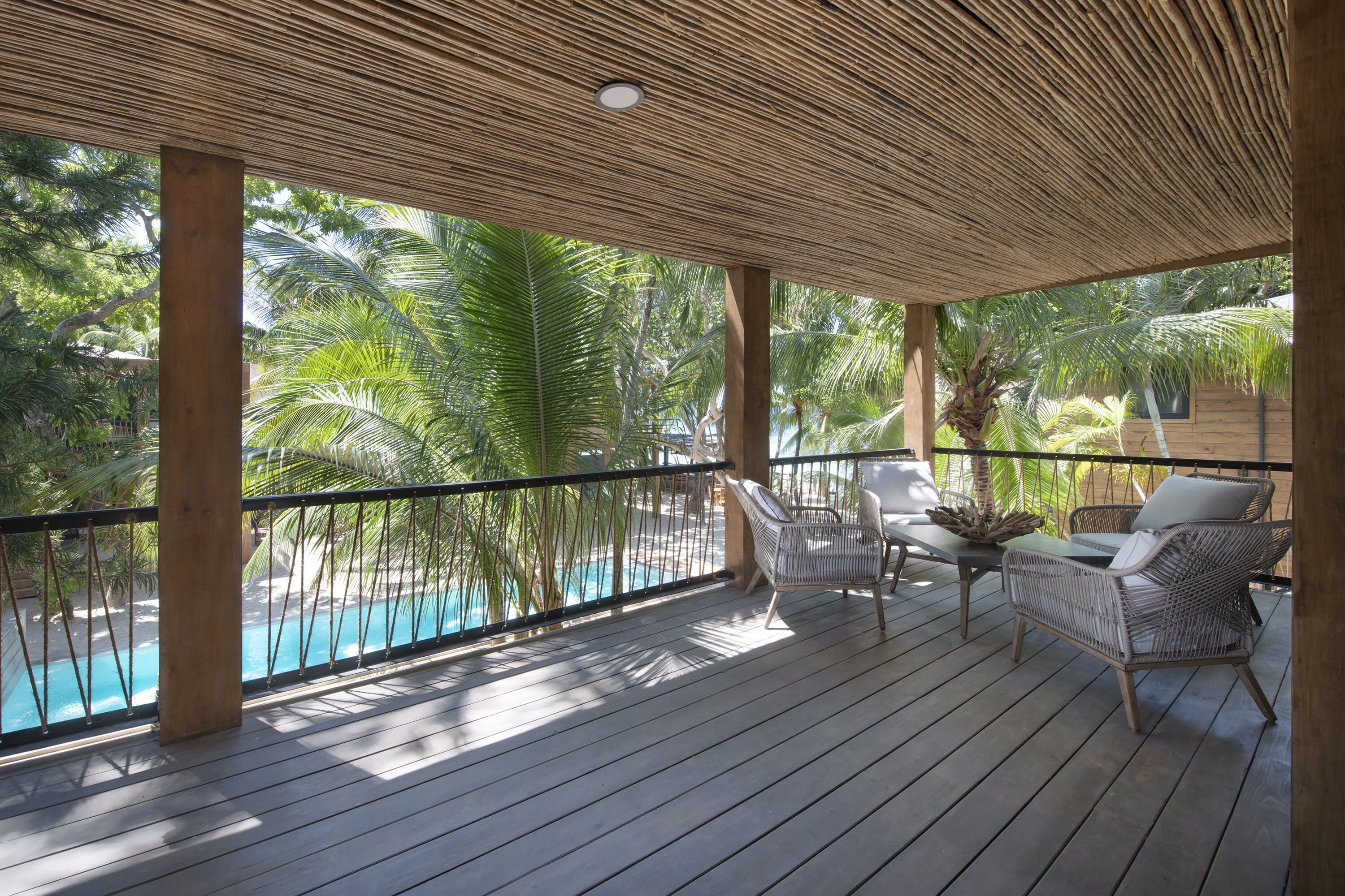 Terraza de madera con vista a la piscina, mobiliario de mimbre y techo de paja.