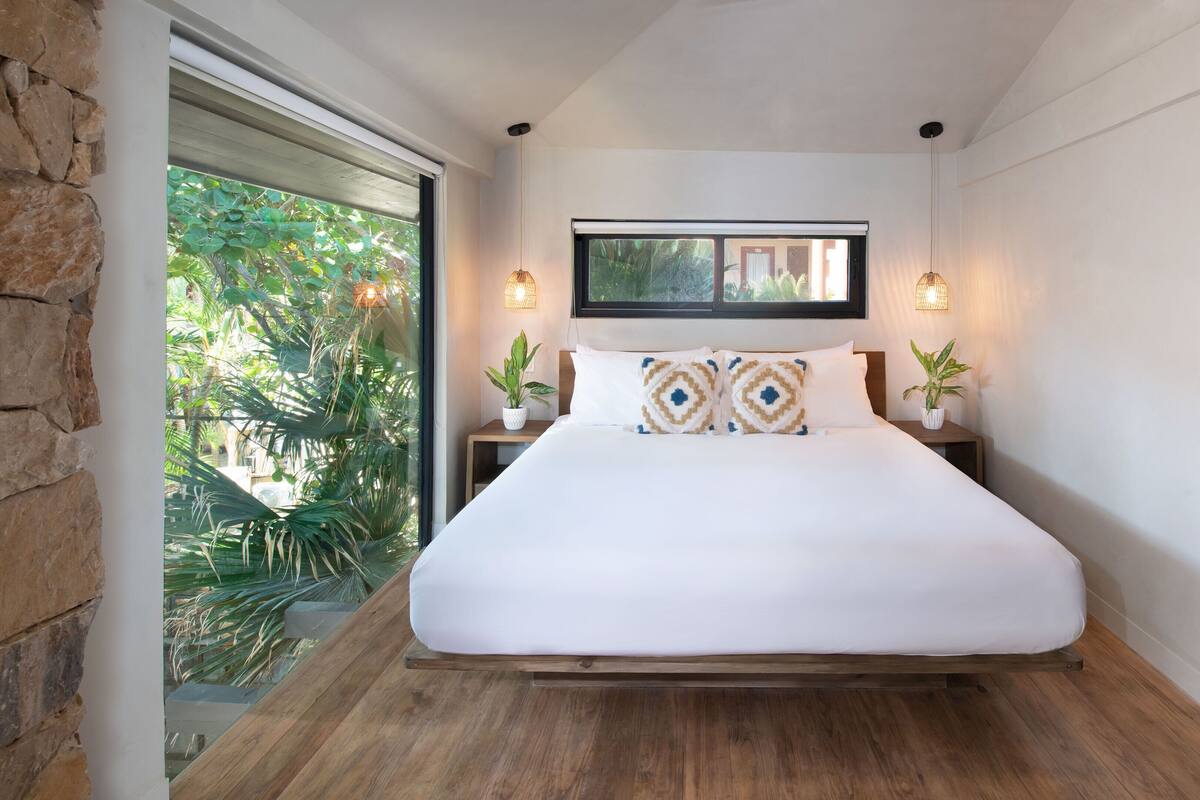 A bedroom with a large bed, two bedside tables with lamps, a window with a view of greenery, and a wooden door.