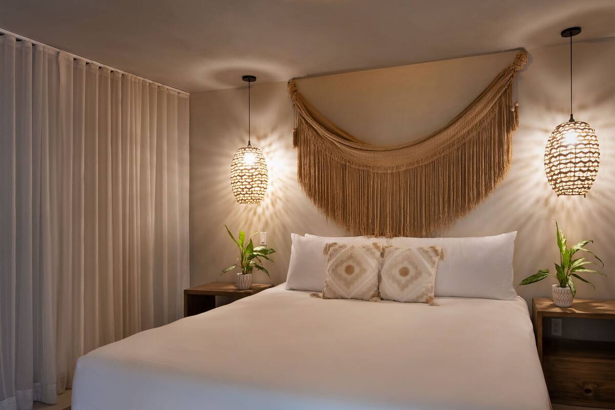 A bedroom with a large bed, two bedside tables with plants, a hanging macramé wall hanging, and a circular ceiling light.