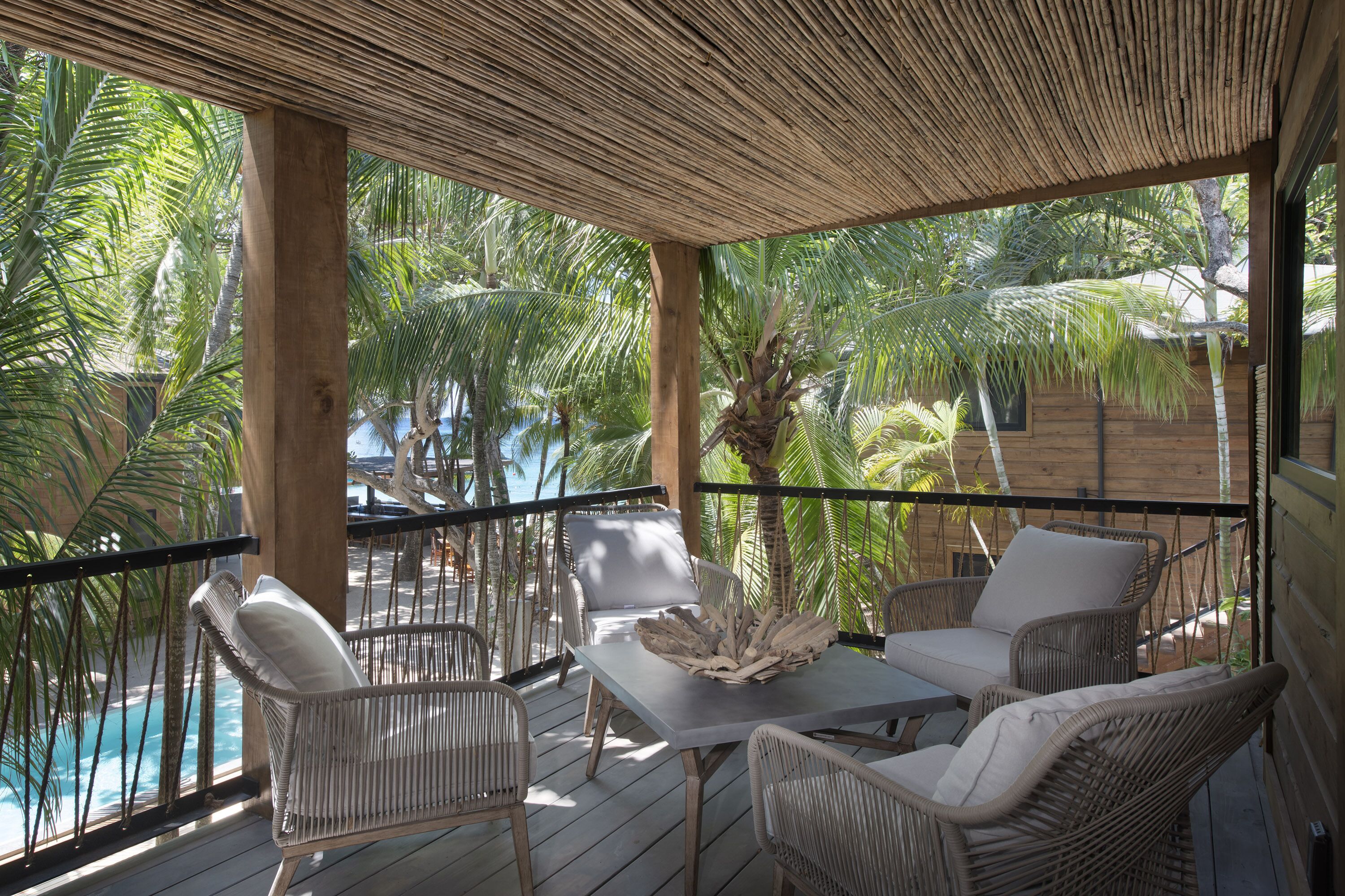 Une terrasse en bois, agrémentée de meubles en osier et d’une table, qui offre une vue sur une piscine et des palmiers.