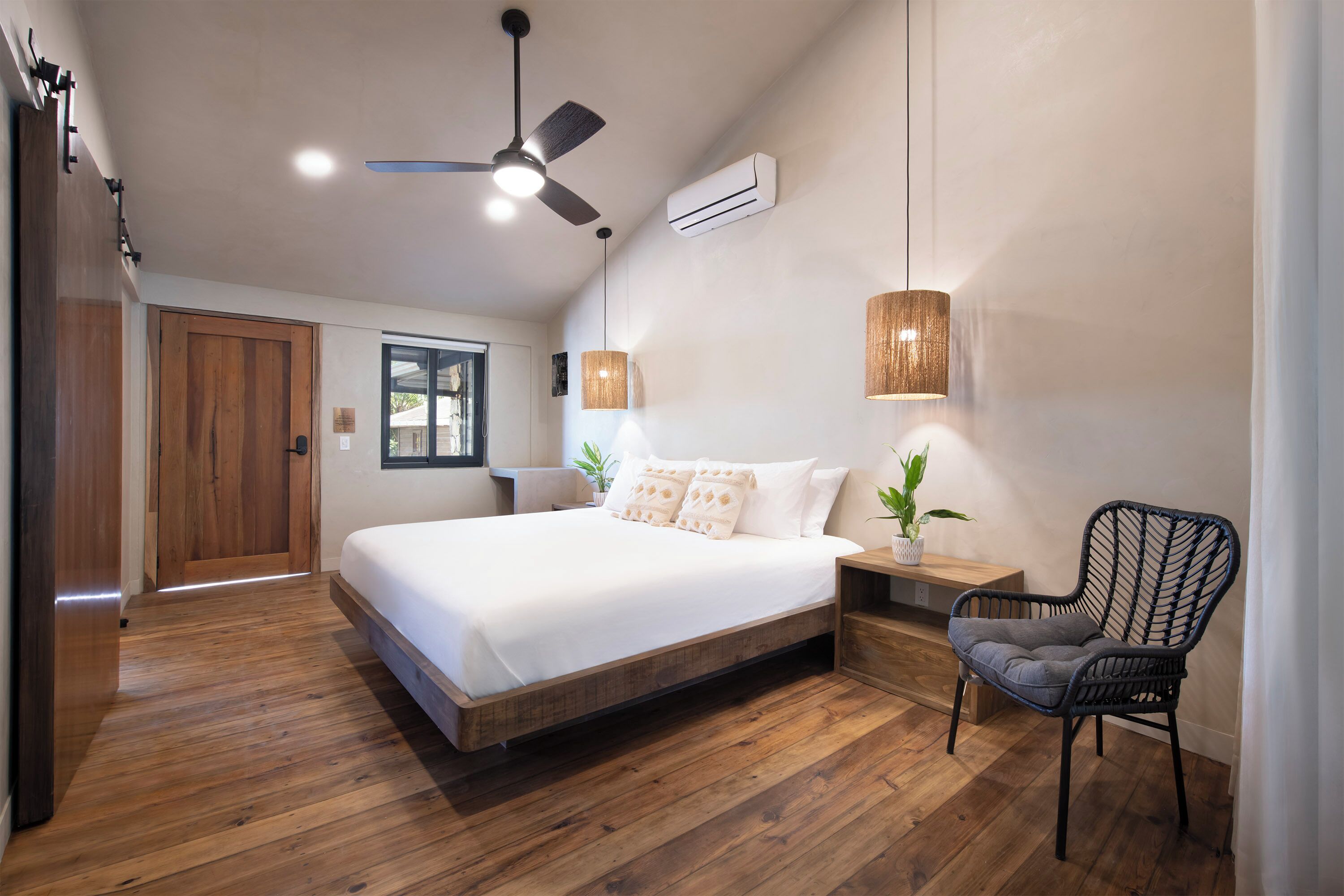 Un dormitorio con piso de madera, una cama con ropa blanca, un ventilador de techo, un aire acondicionado, una ventana y una silla.