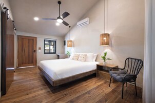 Un dormitorio con piso de madera, una cama con ropa blanca, un ventilador de techo, un aire acondicionado, una ventana y una silla.