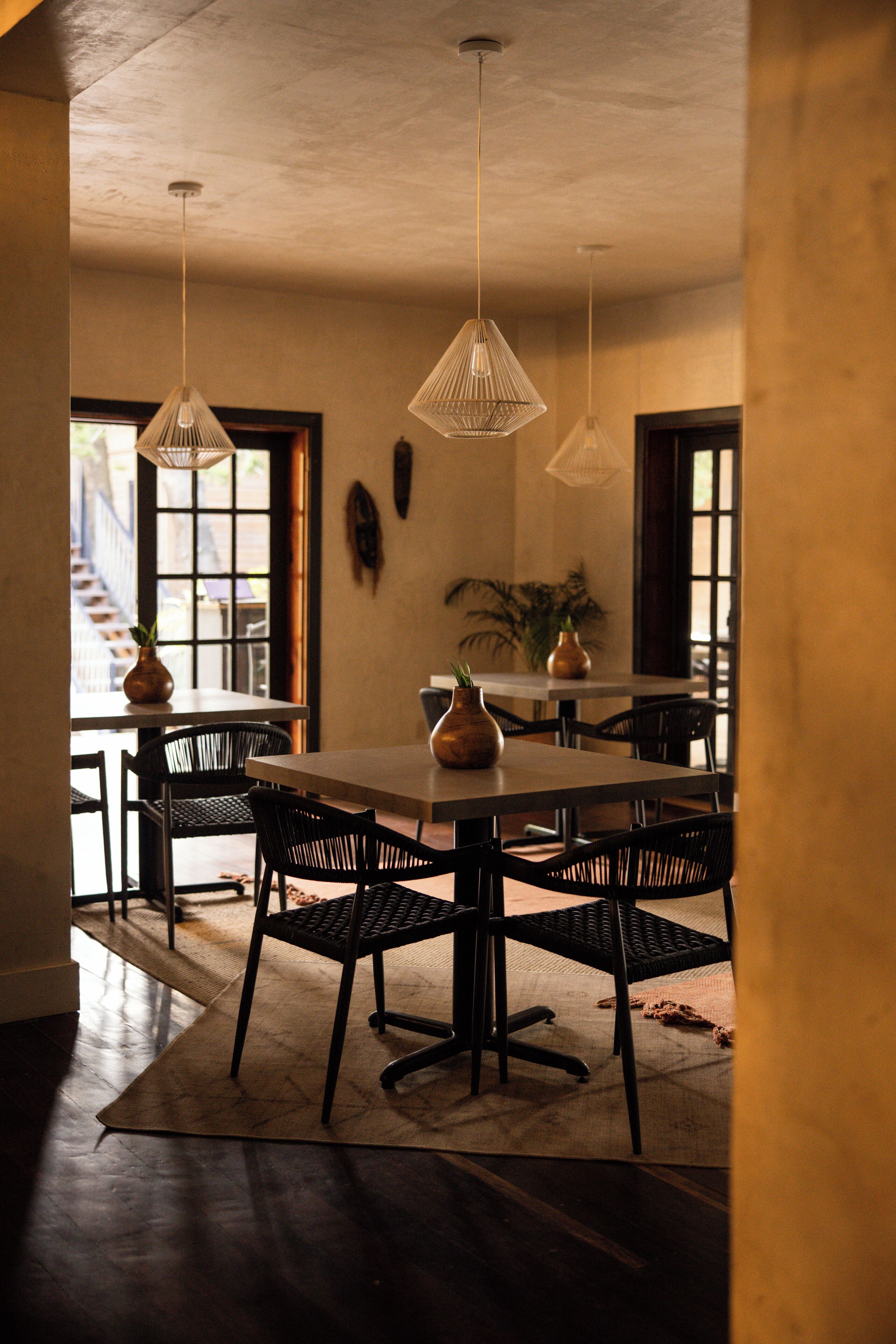 A dining area with a long table, black chairs, hanging pendant lights, and a potted plant.