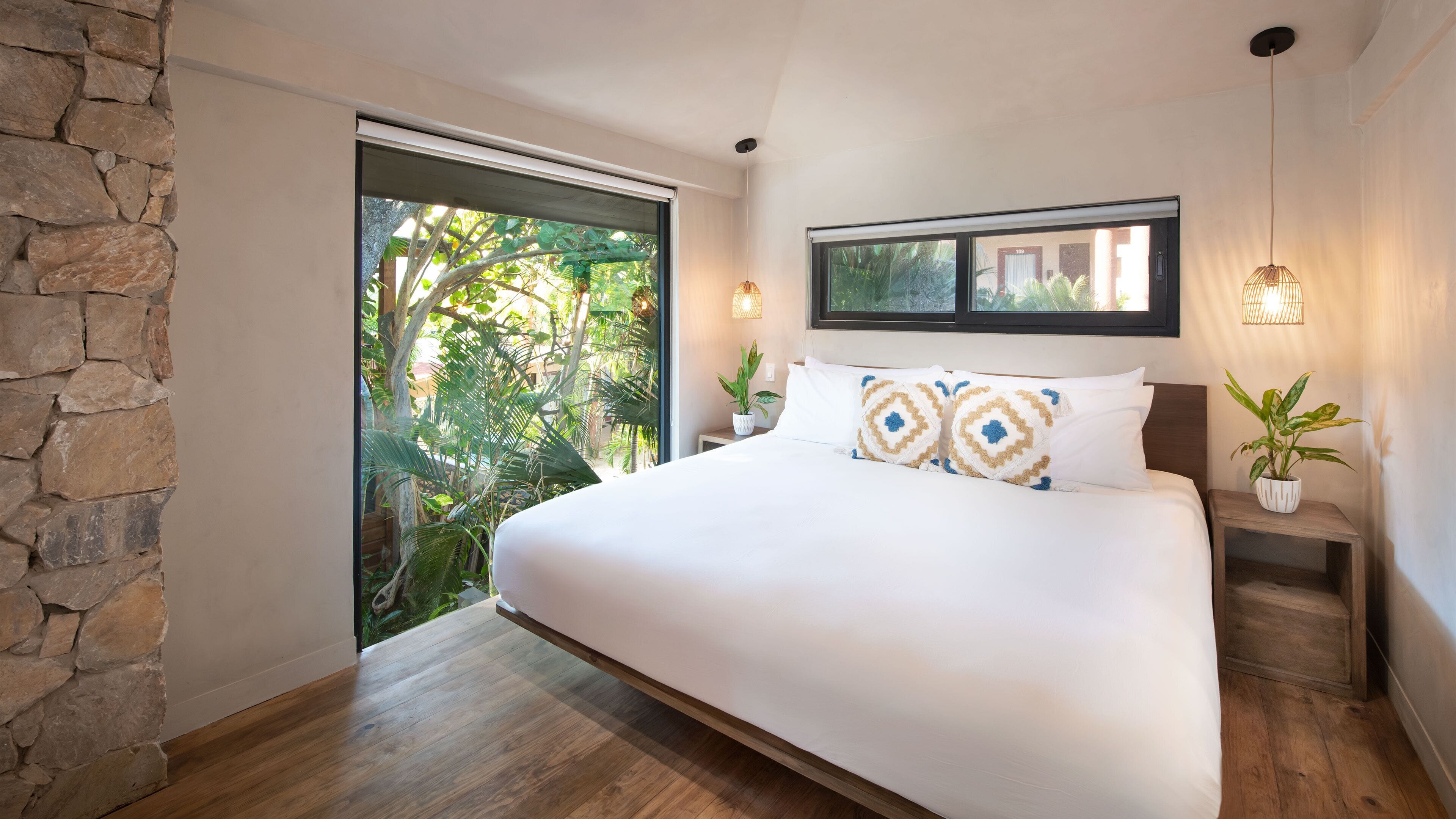 A bedroom with a large bed, stone wall, wooden flooring, a window with a view of greenery, and a small bedside table with a plant.
