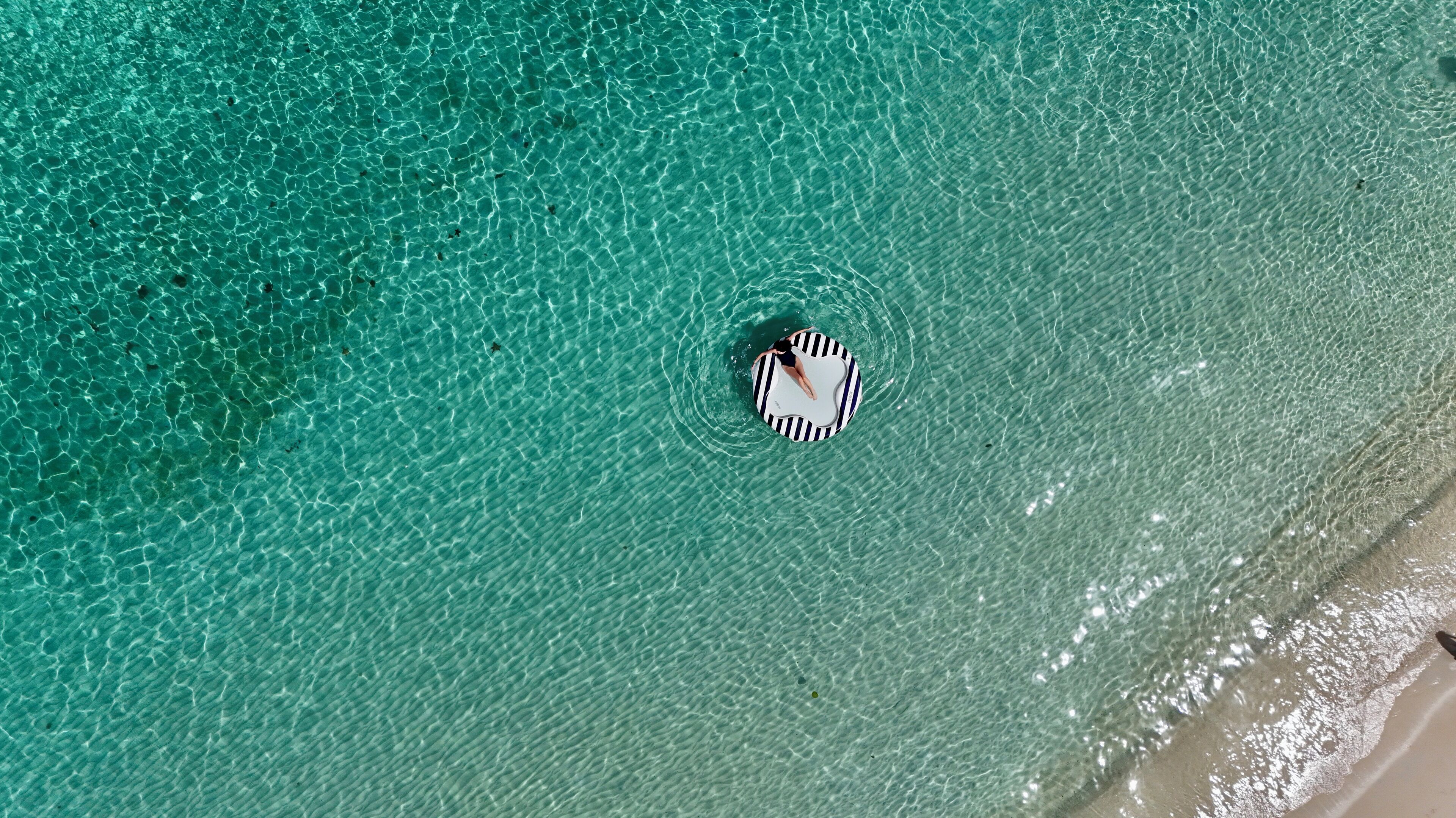 An aerial view of a beach with clear turquoise water and a person on a striped float.