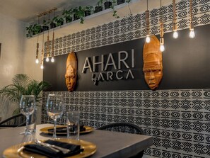A restaurant interior with a patterned wall, wooden head sculptures, hanging light fixtures, and a dining table set with glasses and plates.