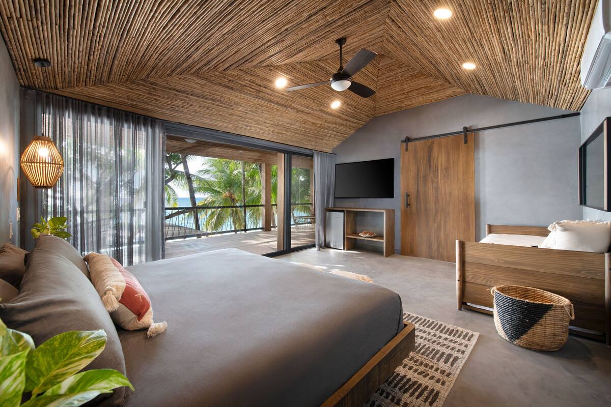 A modern bedroom with a large bed, a TV, and a balcony with a view of palm trees.