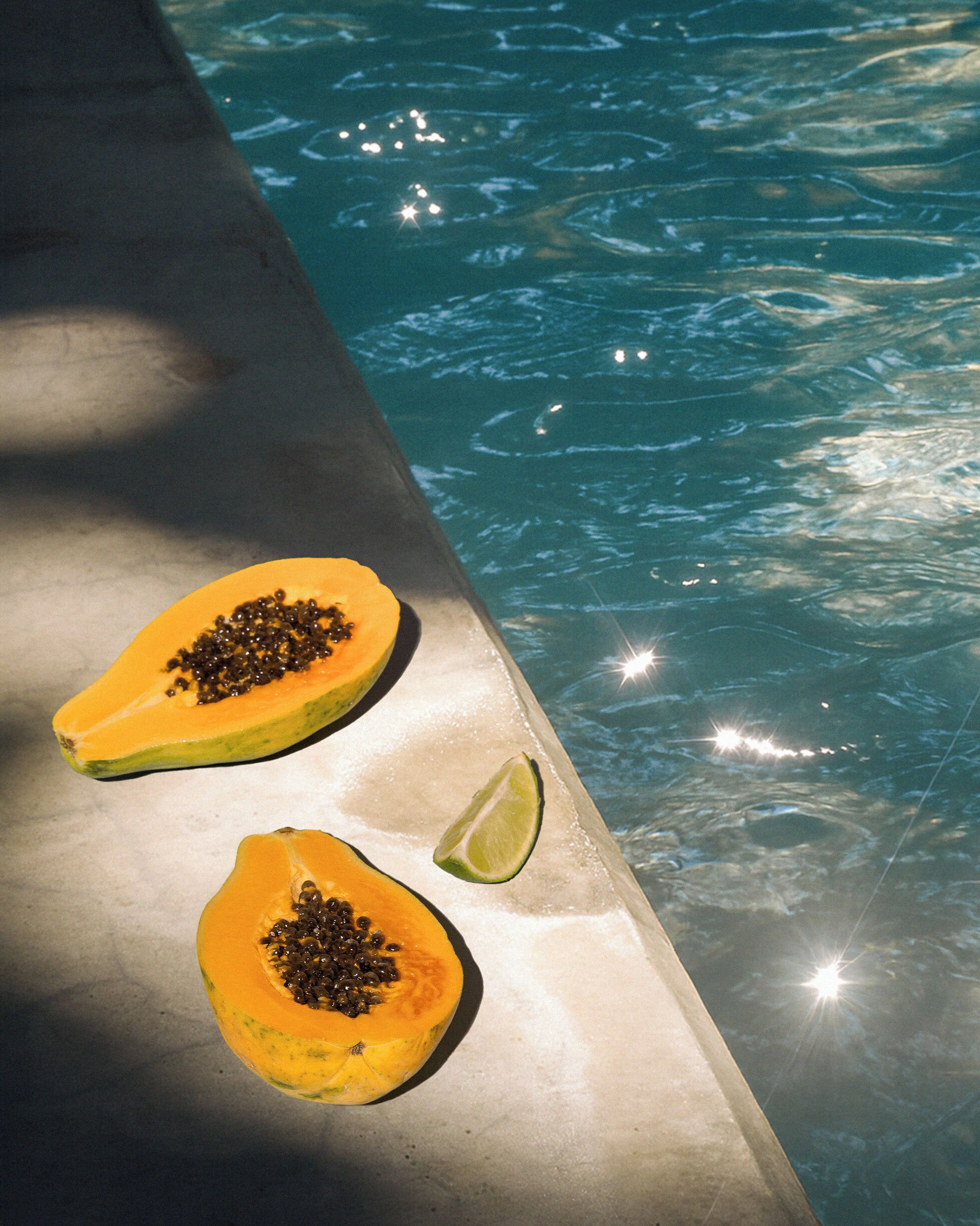 Deux moitiés d’un papaye, avec les graines apparentes, posées sur une surface réfléchissante près d’une piscine.