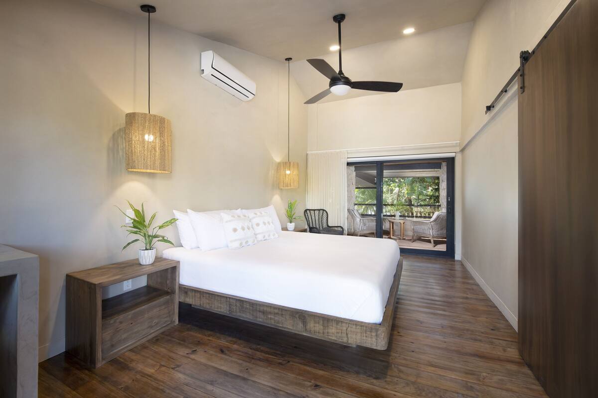 A modern bedroom with a large bed, wooden flooring, a ceiling fan, and a view to an outdoor balcony.