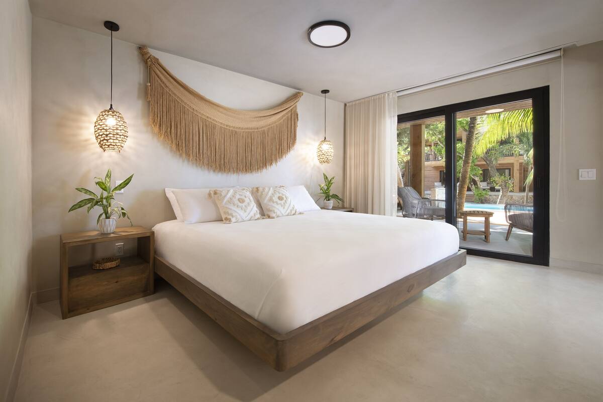 A large bed with a wooden frame, two bedside tables with lamps, a macrame wall hanging, and a view of an outdoor pool area.