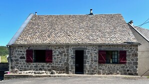A stone building with a sloped roof, two windows with red shutters, and a small door.