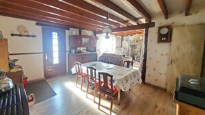 A dining area with a wooden table and chairs, a kitchen in the background, and a clock on the wall.