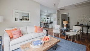 A modern living room with a white sofa, a wooden coffee table, and a dining area with a white chair and a small table. - Beautiful spacious home close to the beach! (Hilton Head Island)