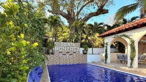 A swimming pool with a sign reading "QUINTA MONARCA" surrounded by lush greenery and a tiled patio area.