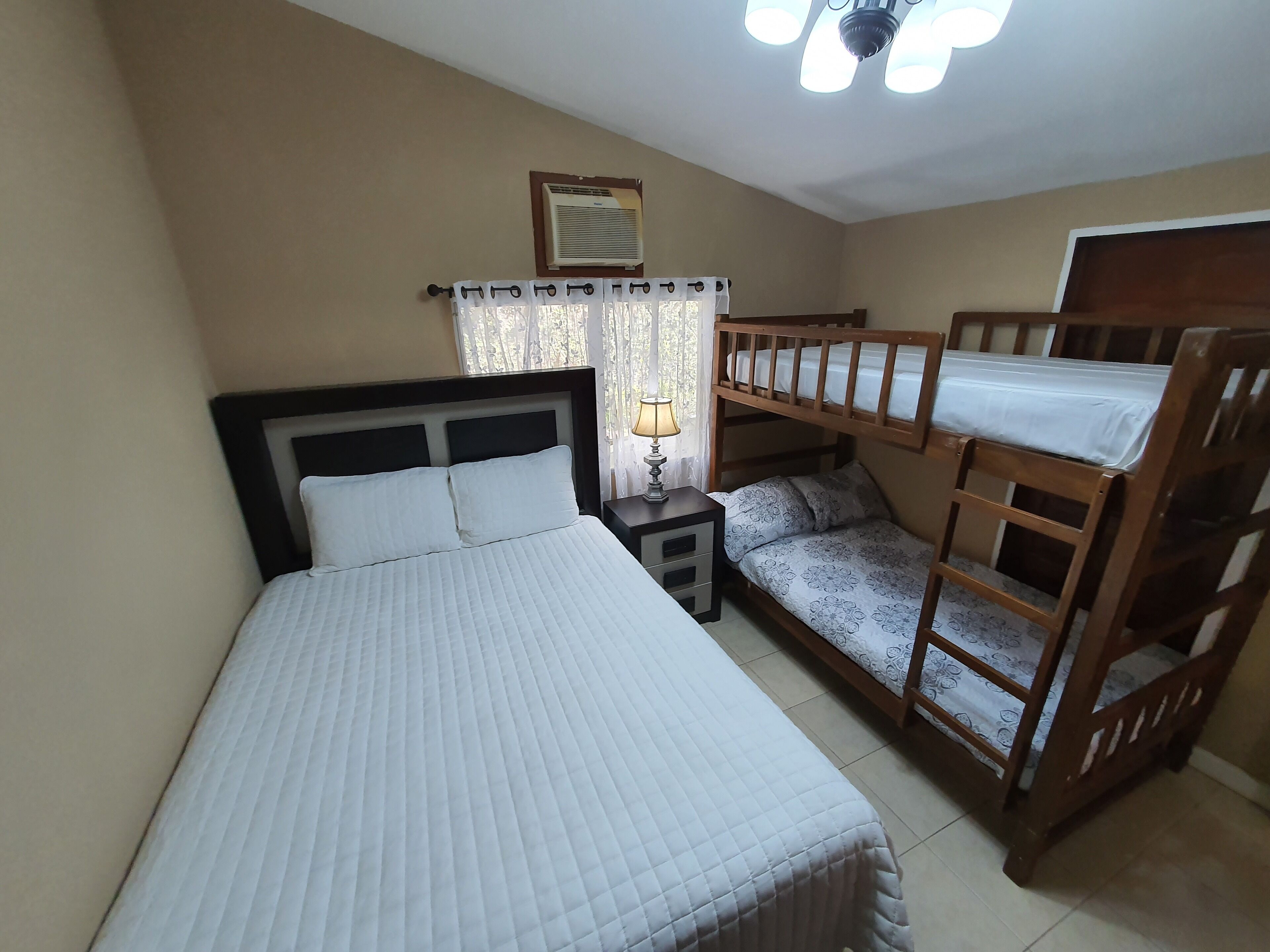 Ein Schlafzimmer mit einem Einzelbett, einem Nachttisch, einer Lampe, einem Fenster mit Vorhängen, einem Deckenventilator und einem Stockbett.