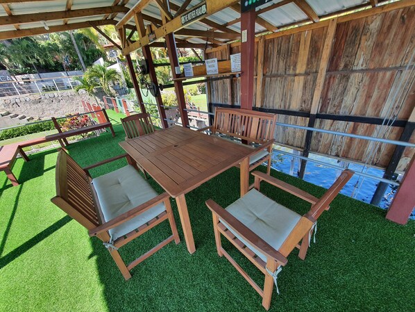 Un conjunto de mesa y sillas de madera al aire libre sobre césped artificial, con una pérgola de madera encima y una vista de un estacionamiento y vegetación al fondo.