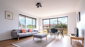 A modern living room with a grey sofa, a small ottoman, a dining table with chairs, a TV on a stand, and large windows offering a view of the outside.