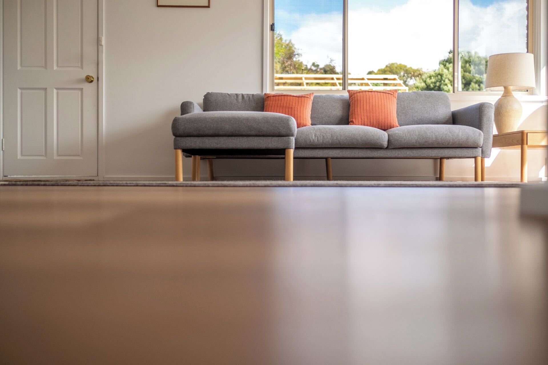 A modern living room with a grey sofa, wooden legs, and a view of the outdoors through large windows.