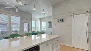 Cocina moderna con una isla grande, muebles blancos y lavavajillas. Hay luces colgantes, un ventilador de techo y un refrigerador.