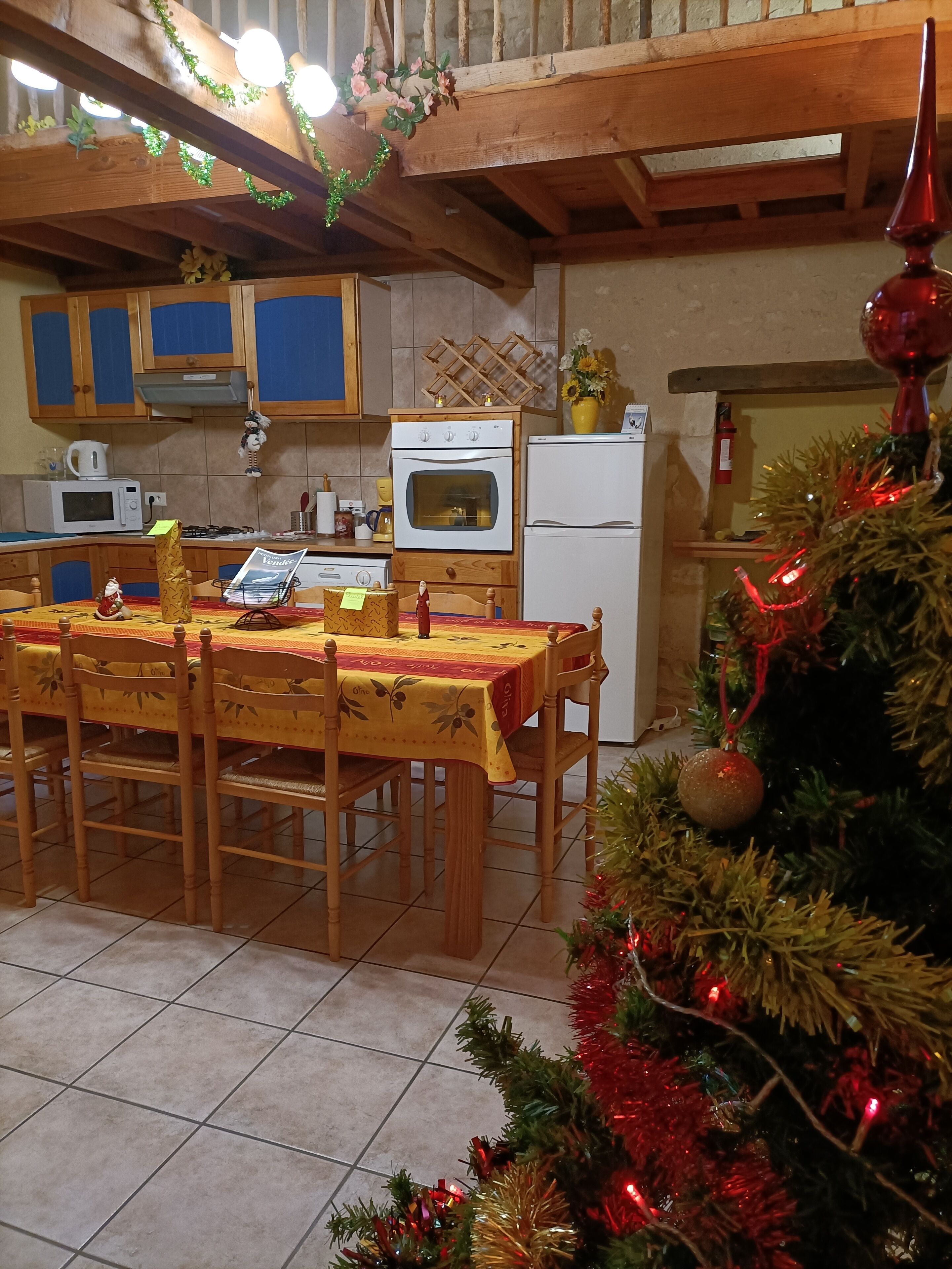 Een keuken met houten kastjes, een eettafel met stoelen, een koelkast en een kerstboom met versieringen.