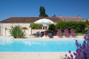 Une piscine entourée de verdure, avec des chaises longues et un parasol, et un bâtiment dont le toit est recouvert de tuiles.