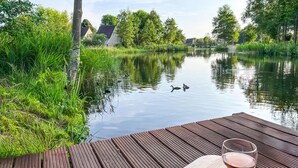Um deck de madeira com uma taça de vinho, com vista para um rio calmo, patos e casas ao fundo.