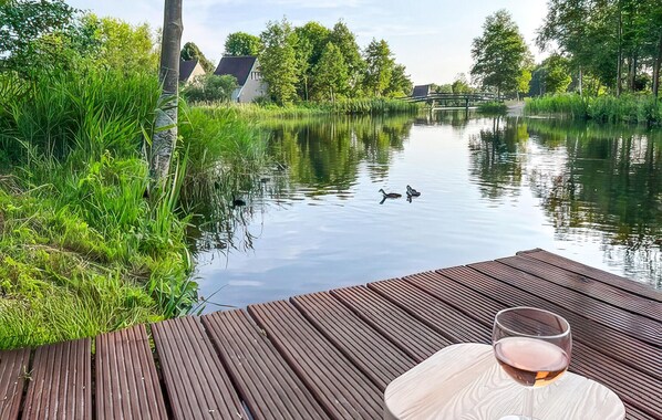 Eine Holzterrasse mit einem Weinglas, mit Blick auf einen ruhigen Fluss, auf dem Enten schwimmen, im Hintergrund sind Häuser zu sehen.