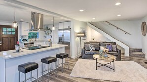 Una cocina moderna con una isla central, taburetes de bar, una zona de comedor con mesa y sillas, un sofá y una escalera.