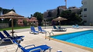 Un’area piscina con poltrone da relax blu e ombrelloni di paglia, circondata da un solarium piastrellato e un edificio in costruzione sullo sfondo.